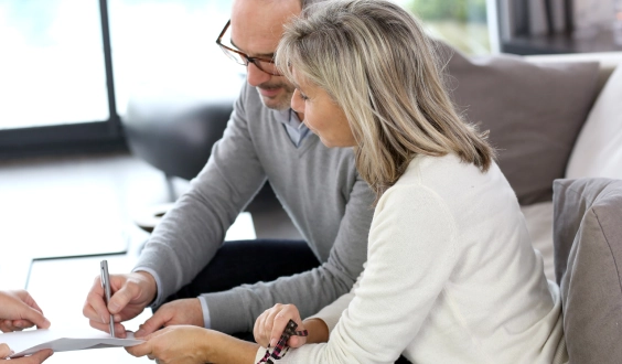 A man and woman sit on a couch together. They are signing a paper with a pen.