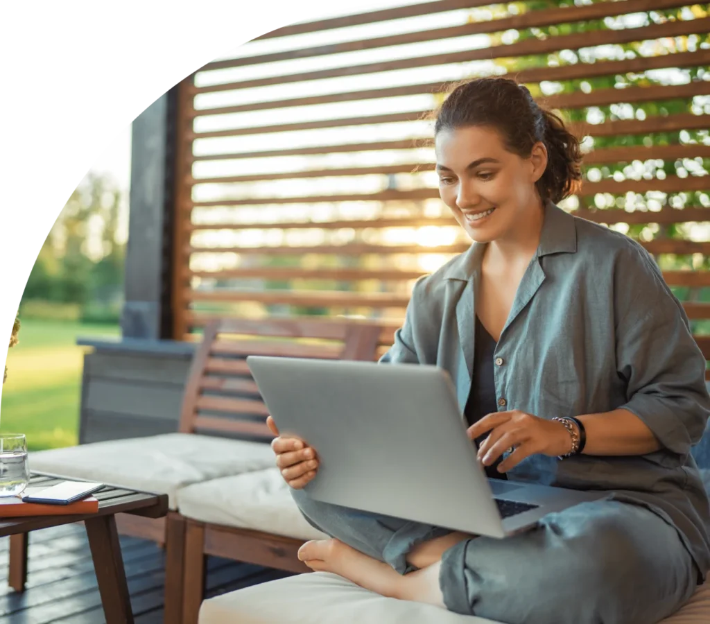 Woman sits in an outdoor living space. She is holding a laptop and smiling.