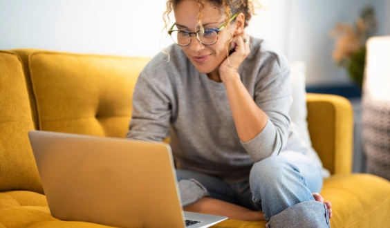 Woman sitting on a yellow couch and looking at a laptop.